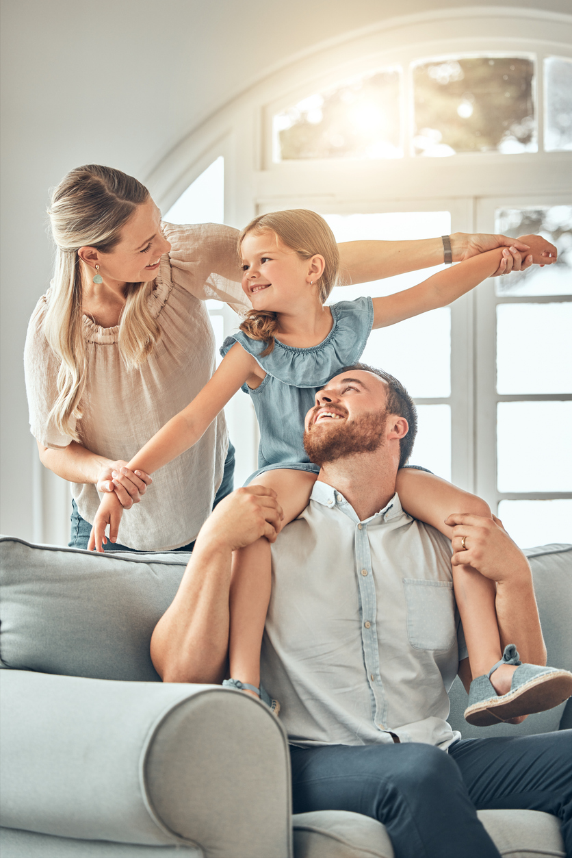 Famille assise dans un salon lumineux, un adulte portant un enfant sur ses épaules. Scène évoquant la protection et la sécurité en assurance pour particuliers.