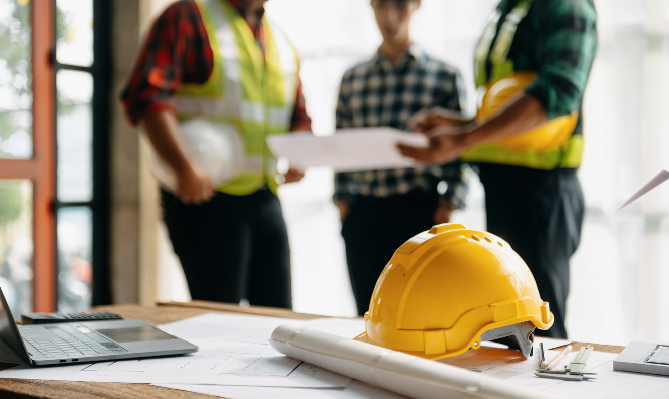 Casque de sécurité posé sur des documents de chantier, avec des intervenants analysant un plan en arrière-plan, en lien avec l’assurance responsabilité des entrepreneurs au Québec.