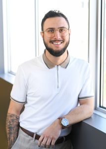 Portrait de Félix Ducharme, conseiller en ressources humaines au Cabinet La Turquoise, debout près d’une fenêtre dans un bureau lumineux, portant des lunettes, un polo clair et une montre.
