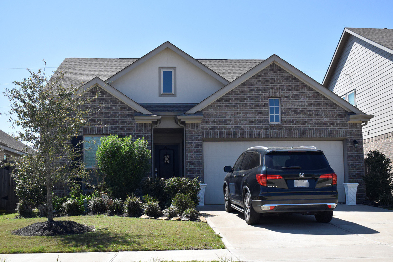 Vue d'une maison unifamiliale en brique avec garage double dans un quartier résidentiel. Un véhicule utilitaire sport noir est stationné dans l'entrée devant la résidence. L'aménagement paysager comprend une pelouse, des arbres et des arbustes, sous un ciel bleu dégagé. Cette scène illustre l'utilisation quotidienne d'un véhicule personnel et son importance pour les déplacements des ménages.