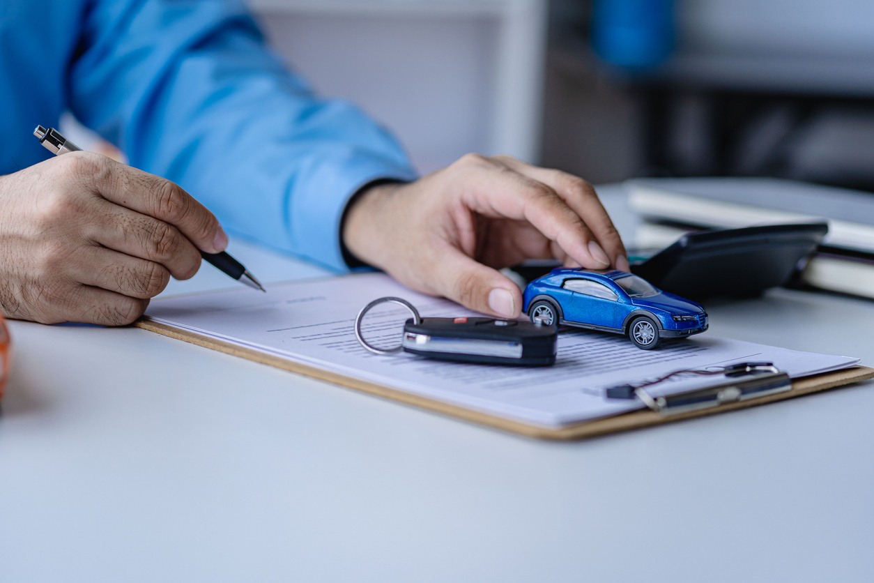 Gros plan d'un bureau où se trouvent des documents, un porte-bloc, des clés de voiture et une miniature de véhicule bleu. Une personne tient un stylo au-dessus des papiers pendant qu'une autre main est posée près du modèle réduit. L'image évoque les démarches administratives liées à l'achat, au financement ou à la protection d'un véhicule.