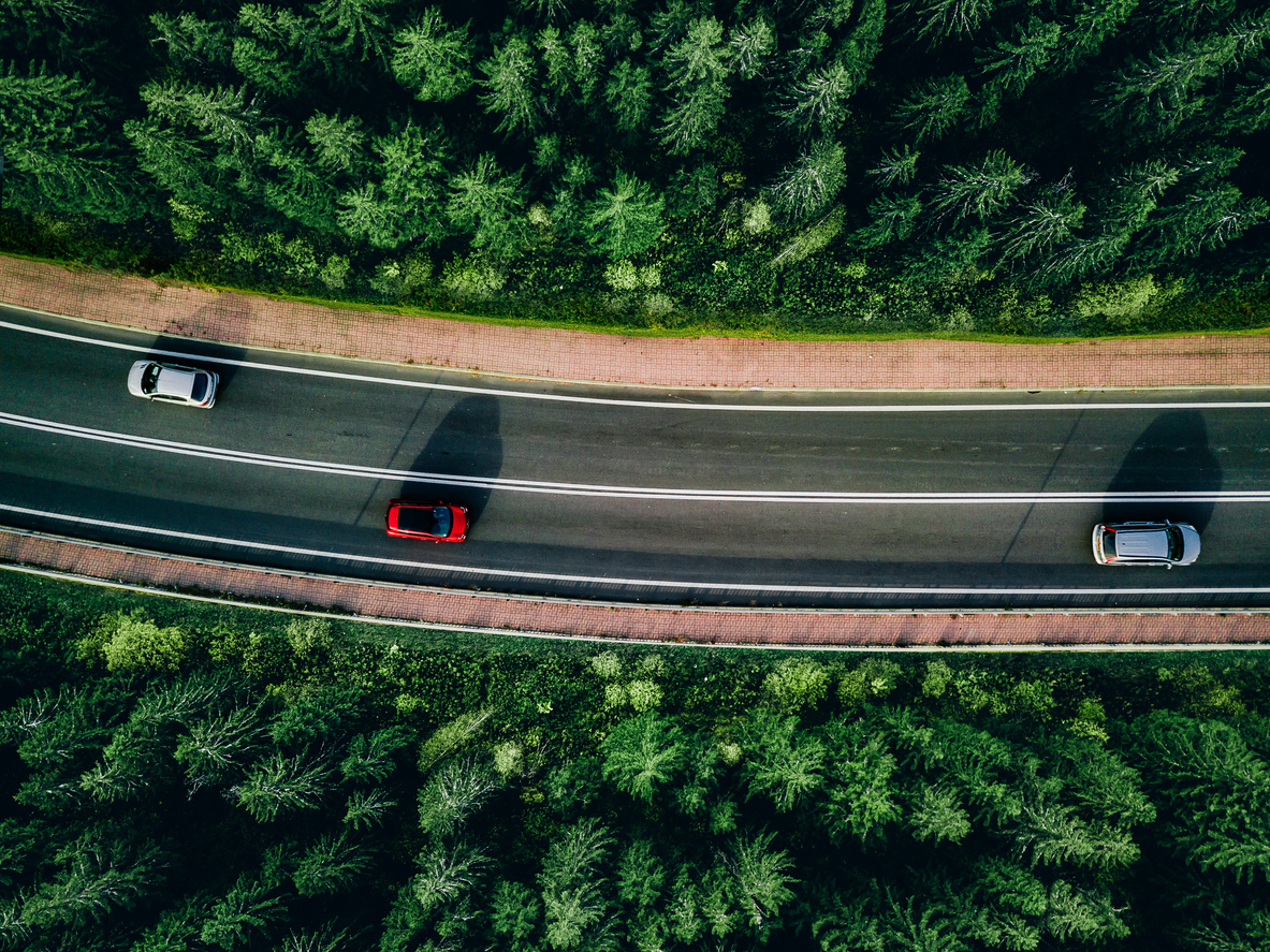 Vue aérienne d'une route traversant une forêt dense composée principalement de conifères. Trois véhicules circulent sur la chaussée dans un environnement naturel verdoyant. L'image met en valeur les déplacements routiers, la mobilité automobile et l'importance de protéger son véhicule pour conserver sa tranquillité d'esprit lors de ses déplacements.