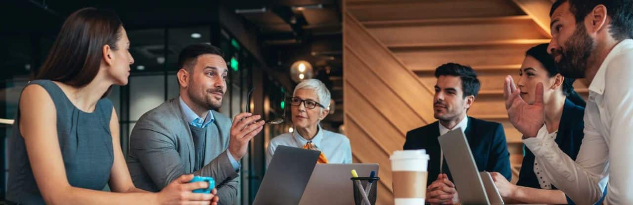 Groupe de cinq personnes réuni autour d’une table dans un environnement professionnel, avec des ordinateurs portables et des cafés, pendant un échange de travail. Groupe de cinq personnes réuni autour d’une table dans un environnement professionnel, avec des ordinateurs portables et des cafés, pendant un échange de travail.
