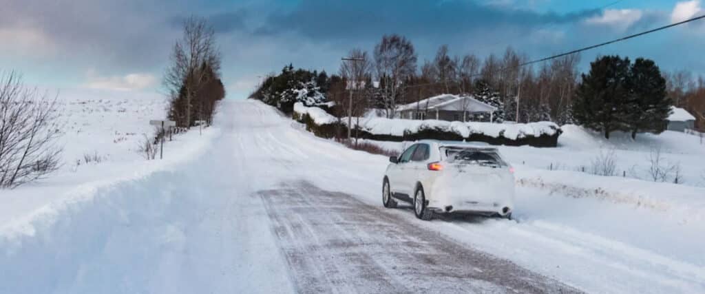 Gros plan sur la roue d’une voiture équipée d’un pneu d’hiver, sur une route partiellement enneigée et glacée. Des traces de pneus sont visibles sur la chaussée, bordée d’une forêt enneigée, tandis que des flocons tombent autour du véhicule. Gros plan sur la roue d’une voiture équipée d’un pneu d’hiver, sur une route partiellement enneigée et glacée. Des traces de pneus sont visibles sur la chaussée, bordée d’une forêt enneigée, tandis que des flocons tombent autour du véhicule.