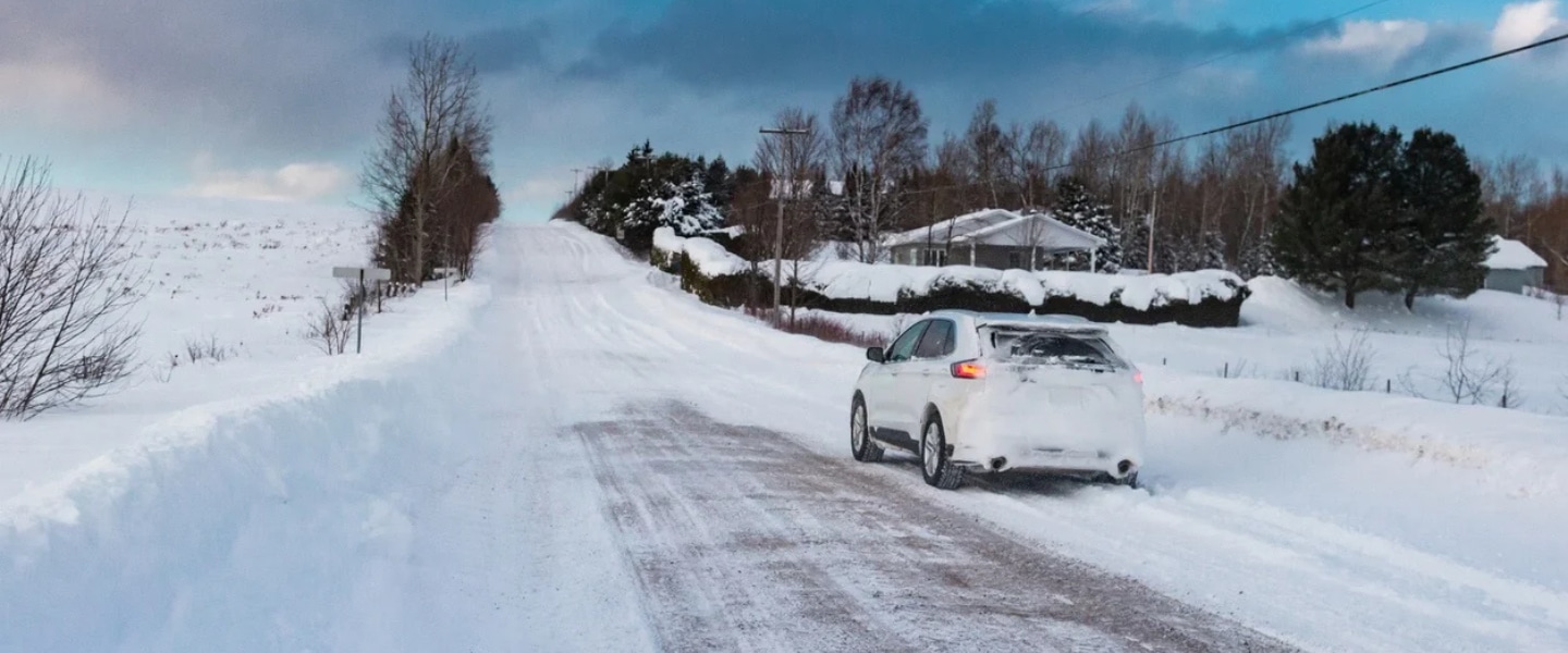 Gros plan sur la roue d’une voiture équipée d’un pneu d’hiver, sur une route partiellement enneigée et glacée. Des traces de pneus sont visibles sur la chaussée, bordée d’une forêt enneigée, tandis que des flocons tombent autour du véhicule. Gros plan sur la roue d’une voiture équipée d’un pneu d’hiver, sur une route partiellement enneigée et glacée. Des traces de pneus sont visibles sur la chaussée, bordée d’une forêt enneigée, tandis que des flocons tombent autour du véhicule.