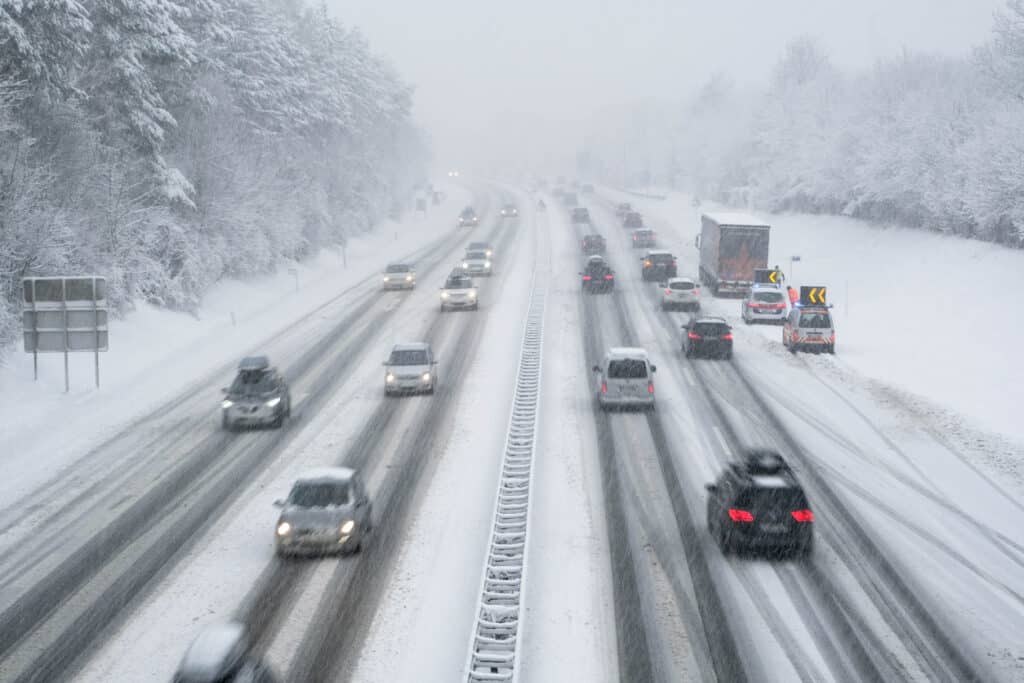 Plusieurs voitures, camionnettes et camions circulent dans les deux directions sur une autoroute enneigée pendant une forte chute de neige. La chaussée présente des traces laissées par les véhicules et l’autoroute est entourée d’arbres entièrement couverts de neige.