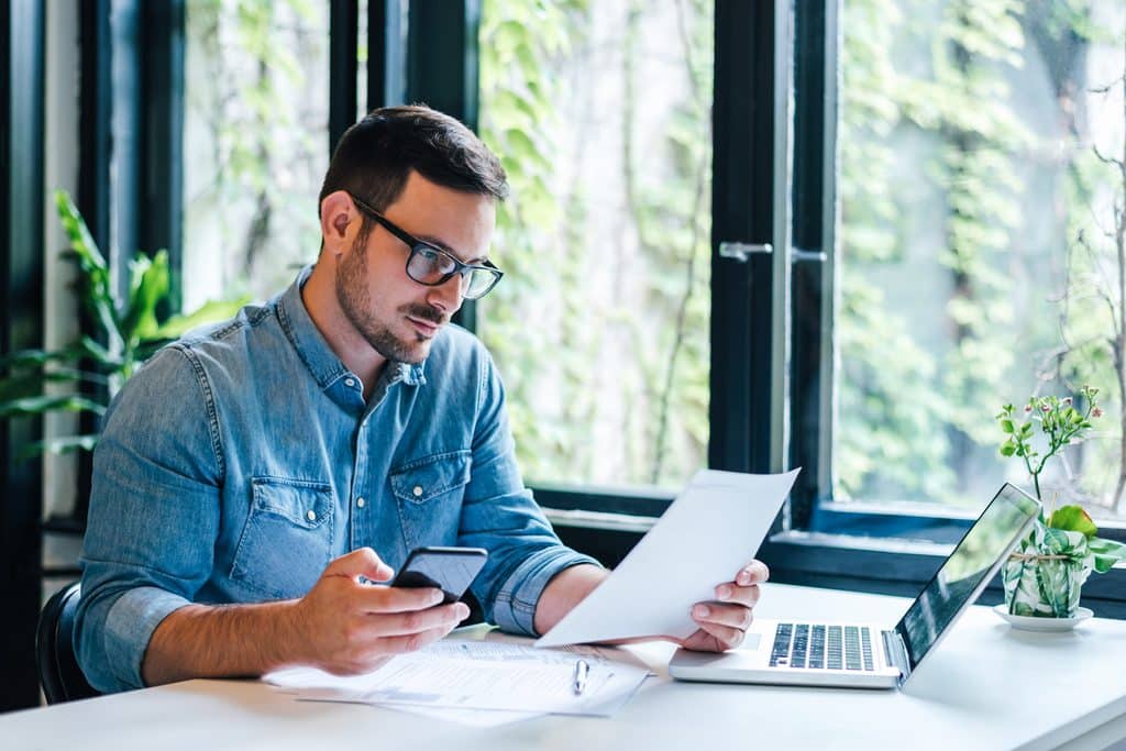 Personne assise à un bureau près de grandes fenêtres, consultant un document papier tout en tenant un téléphone. Un ordinateur portable et plusieurs documents sont disposés sur le bureau.