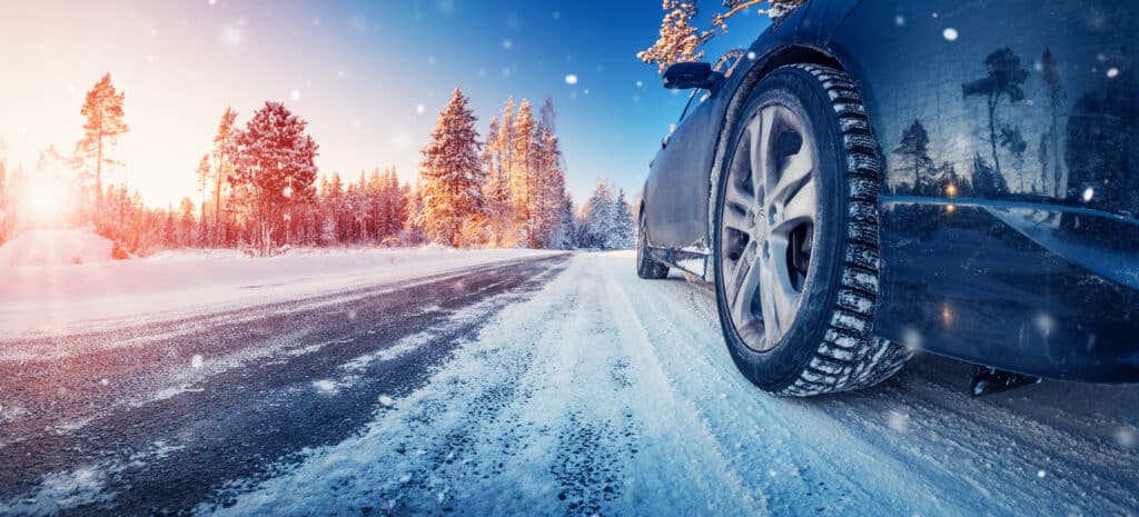 Gros plan sur la roue d’une voiture équipée d’un pneu à bande de roulement prononcée, sur une route partiellement enneigée et glacée. Des traces de pneus traversent la chaussée bordée d’arbres couverts de neige, tandis que des flocons tombent autour du véhicule.