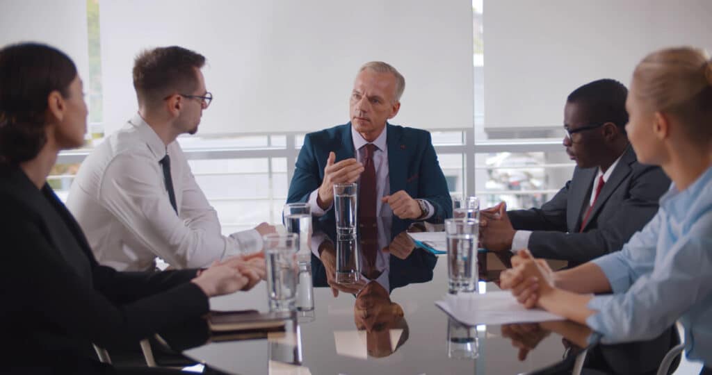 Cinq personnes réunies autour d’une table de conférence dans un bureau. La personne assise au centre prend la parole devant les autres participants, avec des documents et plusieurs verres d’eau disposés sur la table.