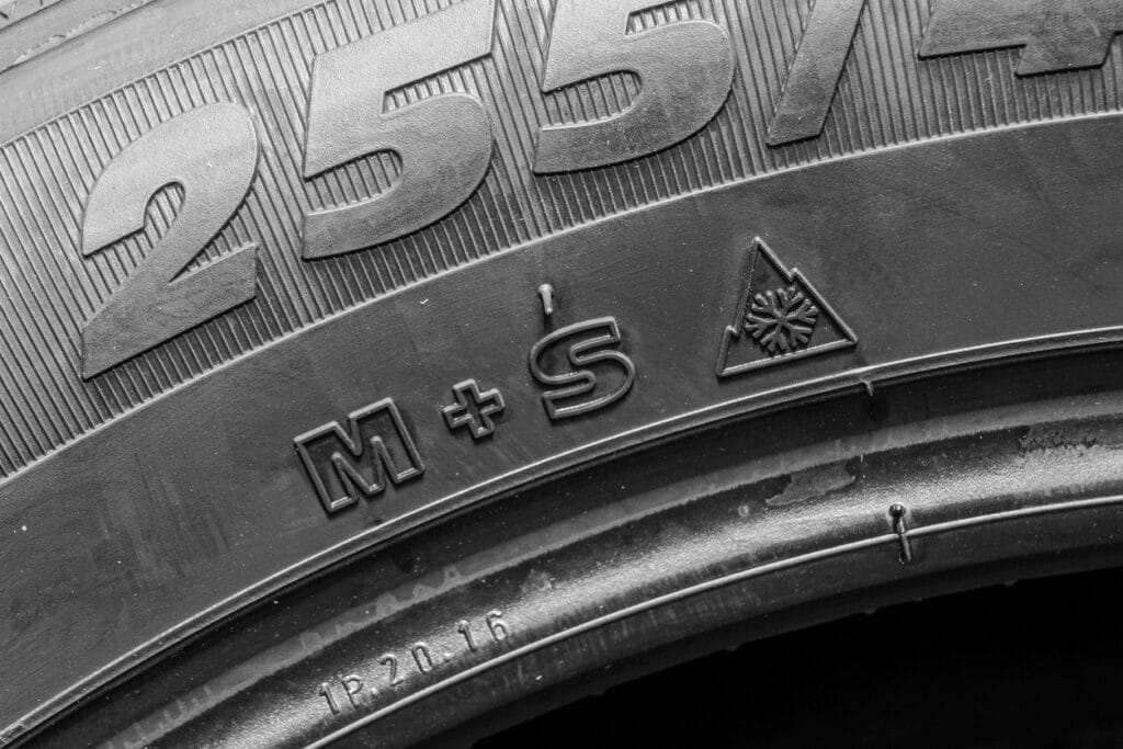 Gros plan en noir et blanc du flanc d’un pneu montrant le marquage « M+S » ainsi que le symbole d’une montagne à trois sommets contenant un flocon. Une partie de la dimension du pneu est également visible au-dessus des deux marquages.