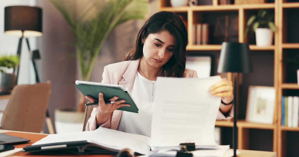 Personne assise à un bureau consultant un document papier tout en tenant une tablette, avec plusieurs dossiers et documents ouverts sur la surface de travail.