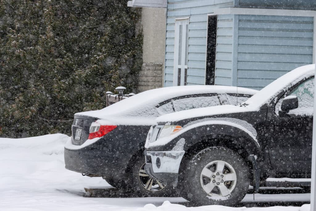 Deux véhicules foncés stationnés côte à côte devant une résidence pendant une chute de neige. Les carrosseries, les vitres et les pneus sont partiellement couverts de neige, tandis qu’une épaisse couche de neige recouvre le sol autour des véhicules.